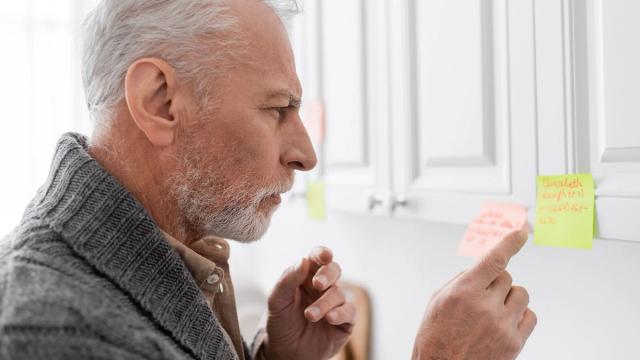 An older man with a gray beard points at a pink sticky note on white kitchen cabinets.