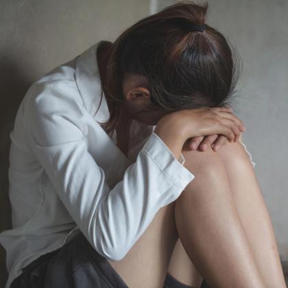Woman sitting on the floor with her head down, resting on her knees, appearing distressed or upset, wearing a white shirt and dark shorts.