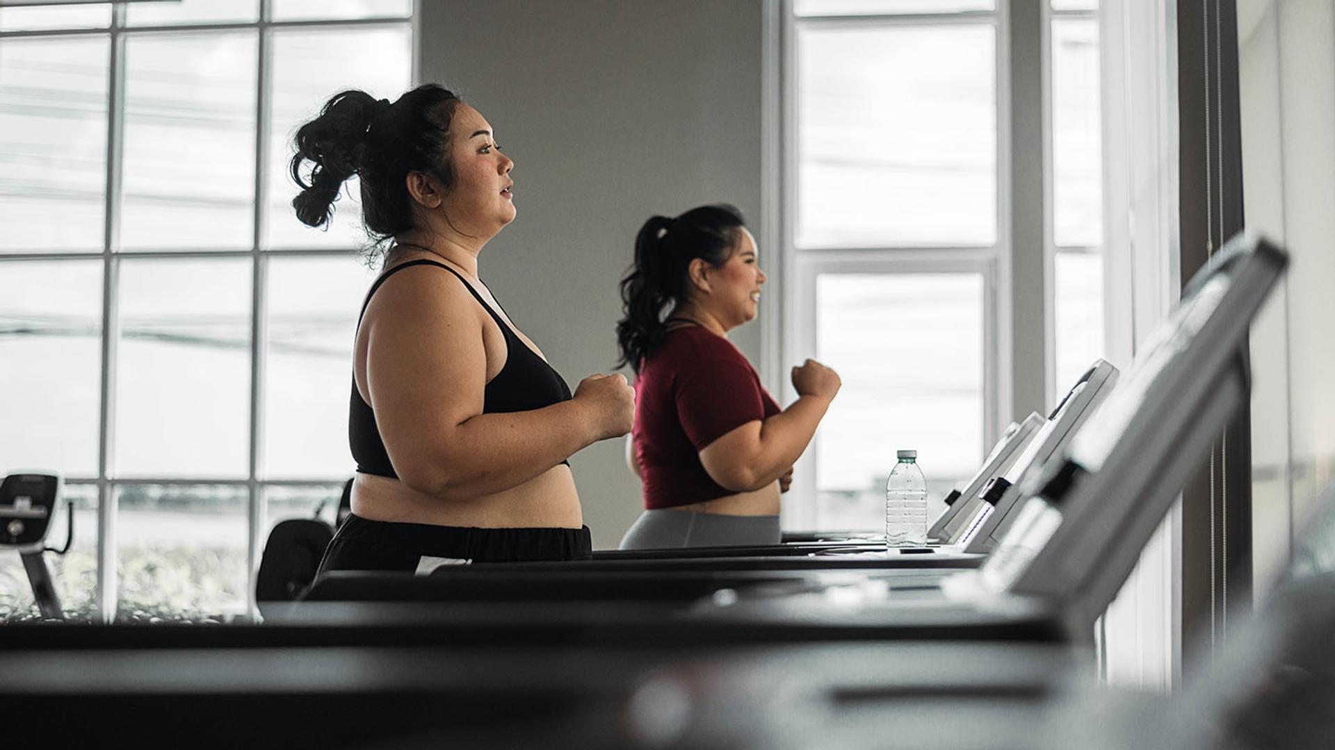 Two plus-size women running on treadmills in a gym.