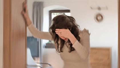 Woman with hand on head, leaning on a door frame, experiencing vertigo in a blurred room.