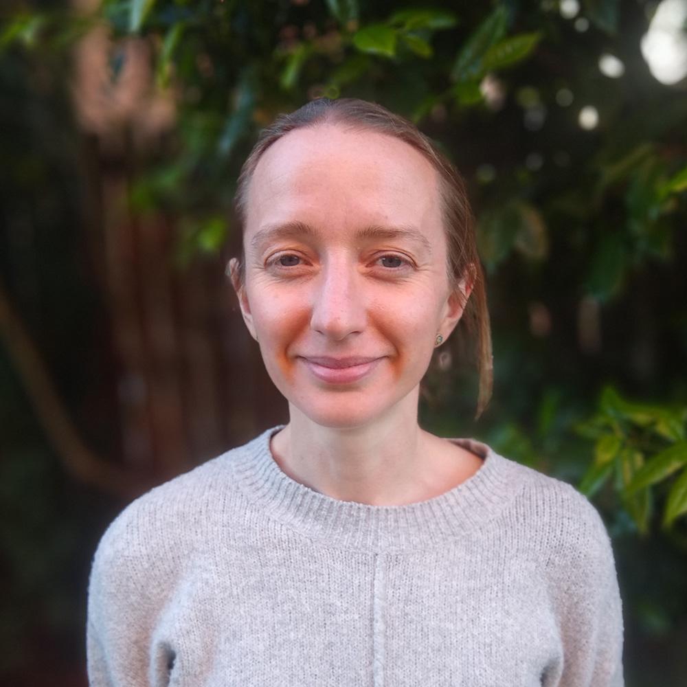 Portrait of a smiling woman with light brown hair wearing a grey sweater, standing in front of lush green foliage.