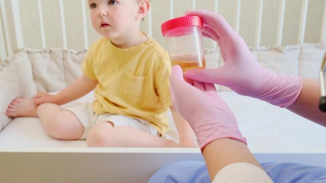 Child in a crib looking at gloved hands holding a urine sample cup.