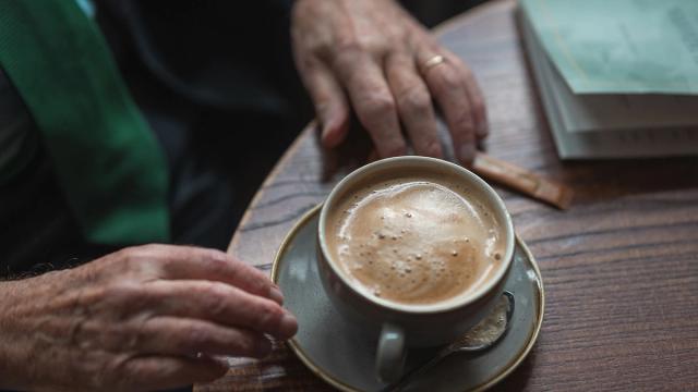 A cup of foamy coffee on a wooden table, with an older person's hands and a book.