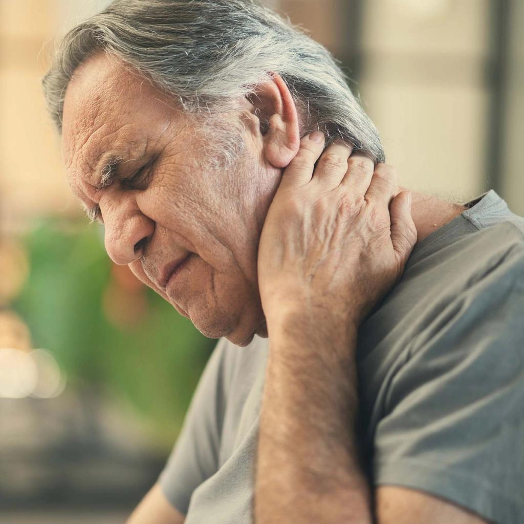 An older man with grey hair holds his neck with one hand, his eyes closed in pain.