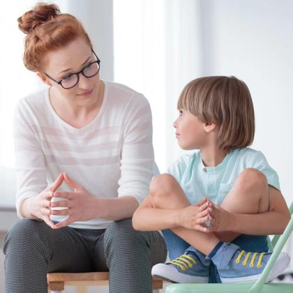 Woman with glasses talking to young boy sitting on a chair in a bright room