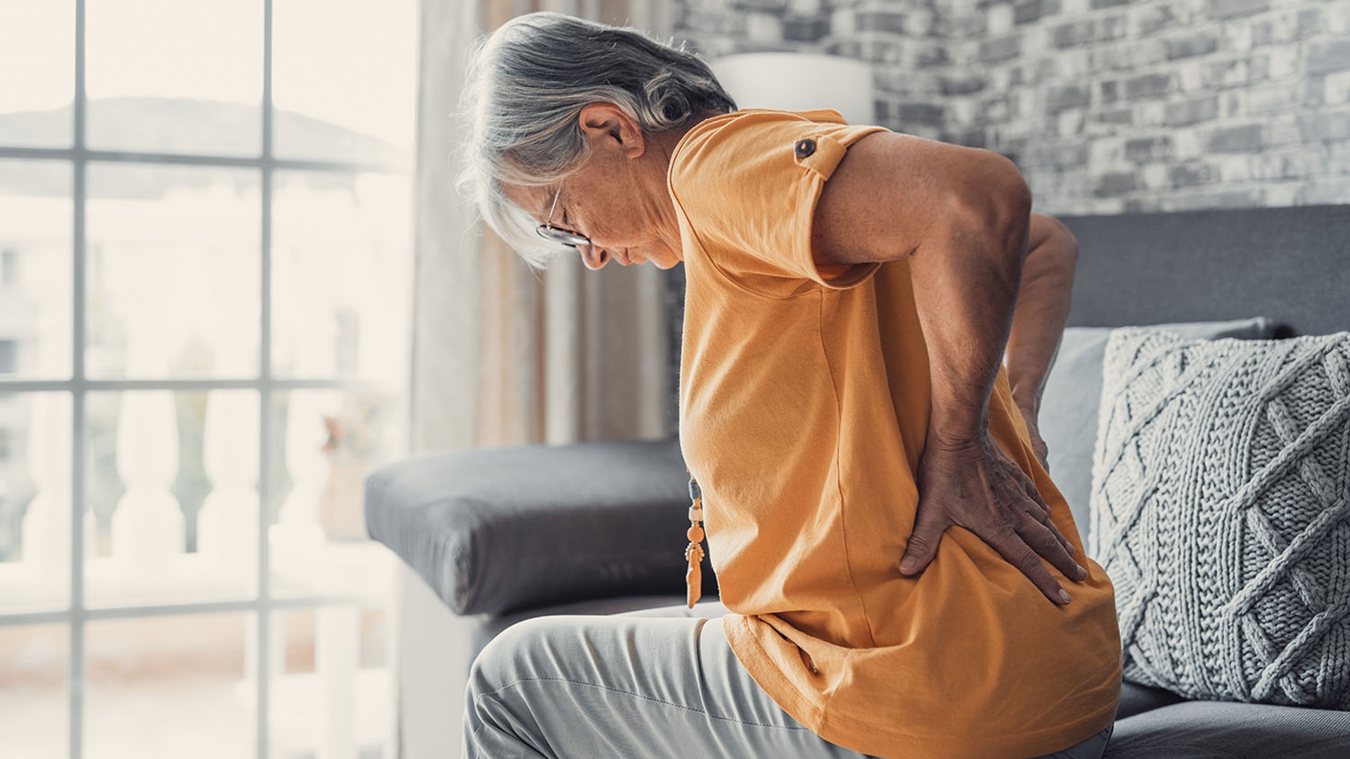 Elderly woman sitting on a sofa clutching her lower back in pain.
