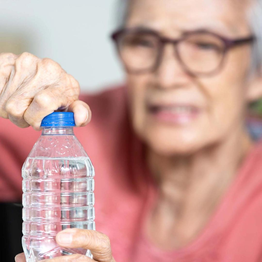 An elderly person's hands struggle to open a water bottle, with their blurred face showing effort.