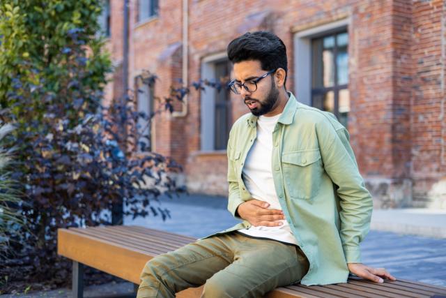 A young man sits on a bench outdoors, clutching his stomach in discomfort.