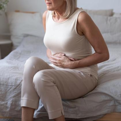 Senior woman sitting on bed holding stomach, possibly experiencing pain or discomfort.
