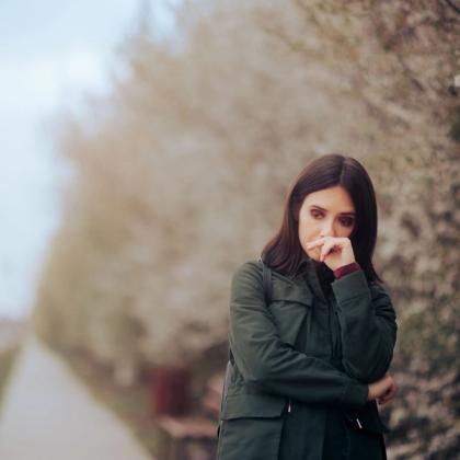 Woman in a green jacket leaning thoughtfully on her hand, standing on a tree-lined path.