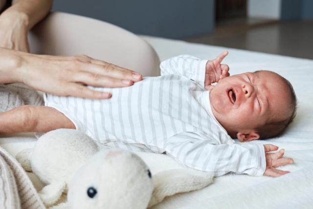 An adult's hand gently rests on a crying newborn baby's stomach.