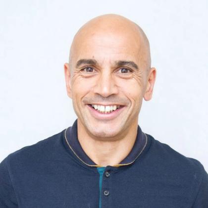 Bald man with a friendly smile wearing a dark collared shirt against a plain light background.