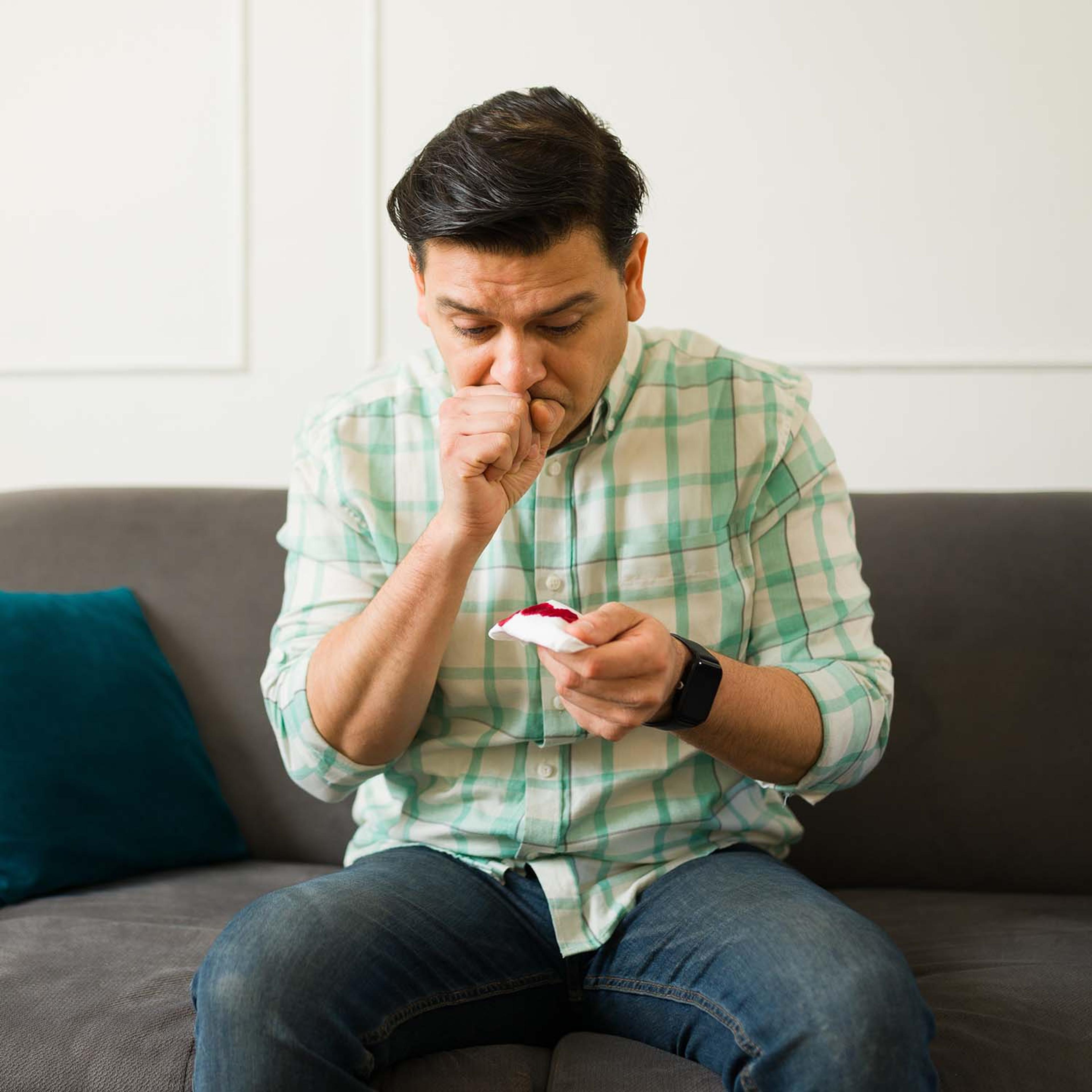 Man on couch holding a blood-stained tissue and coughing.