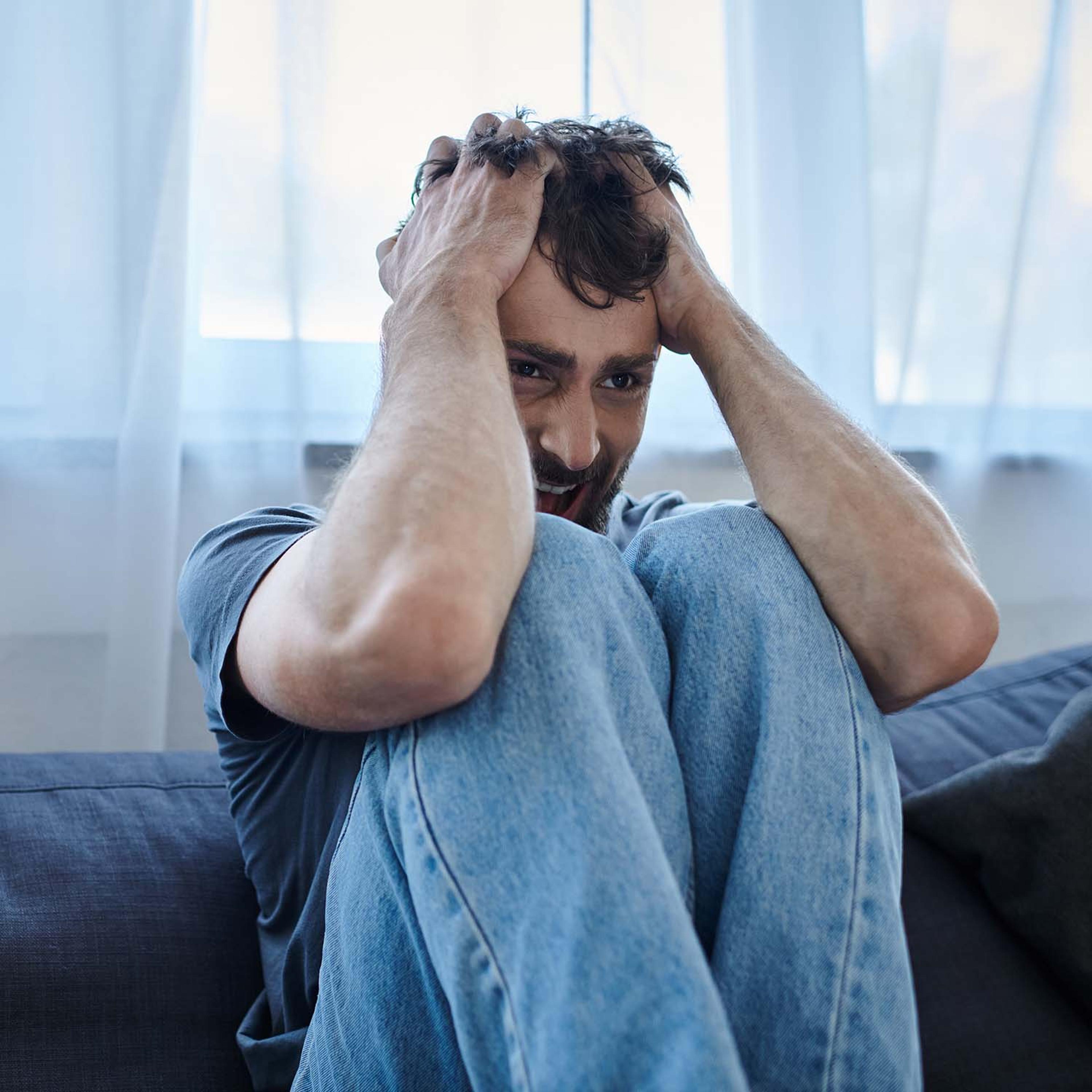 A man in distress sits on a couch, clutching his head in his hands.