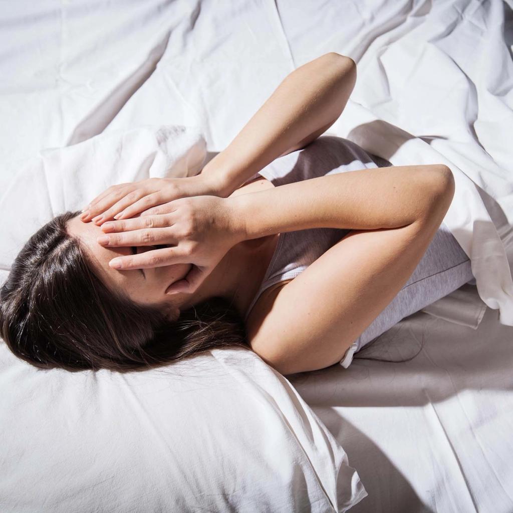 A woman lies in bed, covering her eyes from the bright light.