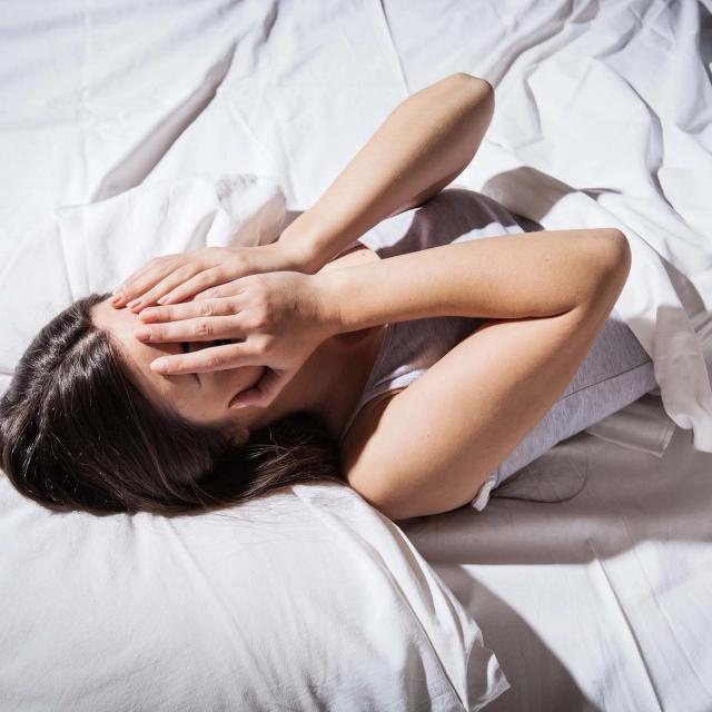 A woman lies in bed, covering her eyes from the bright light.