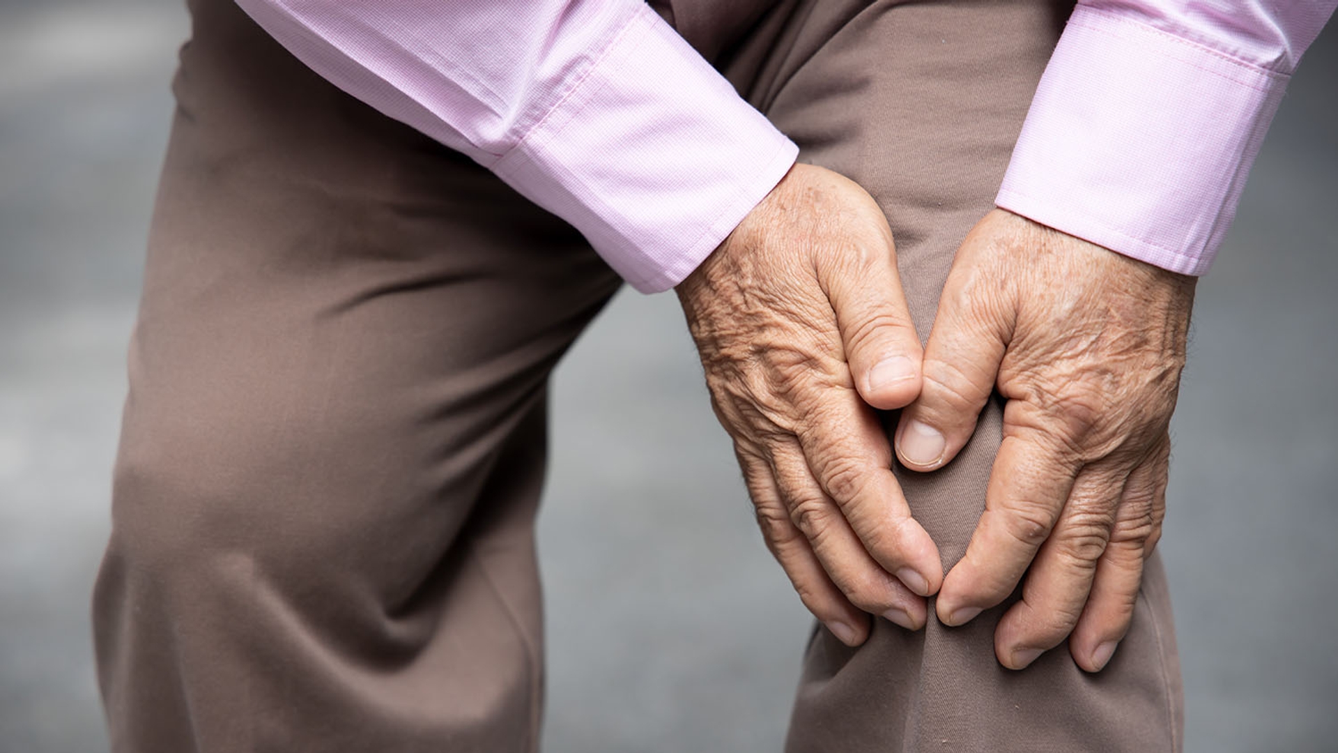 Close-up of an elderly person's hands clasped behind their back, wearing a pink shirt and brown trousers.