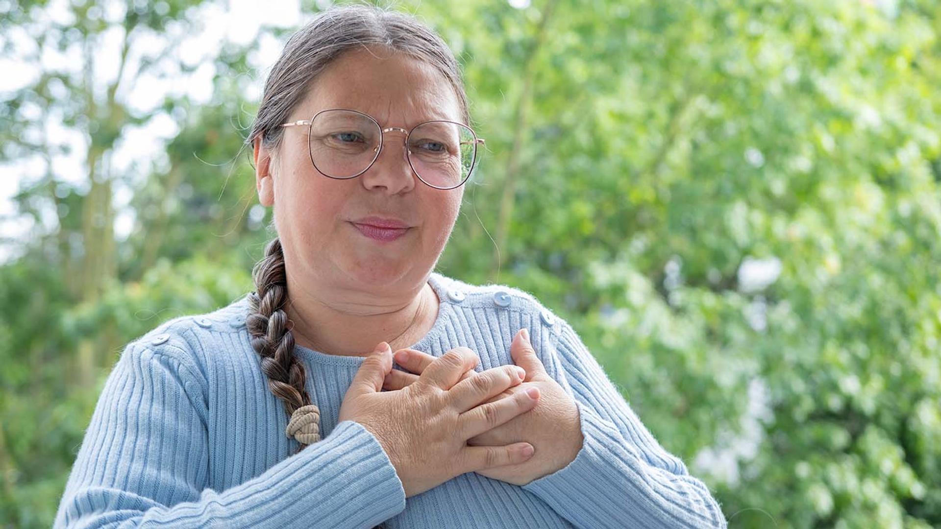 A woman with glasses clutches her chest with an expression of pain or discomfort.