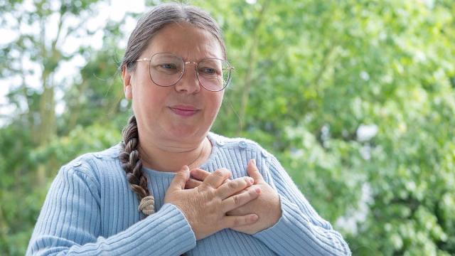 A woman with glasses clutches her chest with an expression of pain or discomfort.