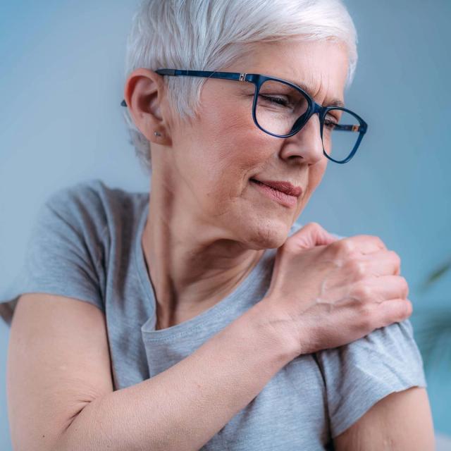 An older woman with grey hair and glasses holds her shoulder in pain.