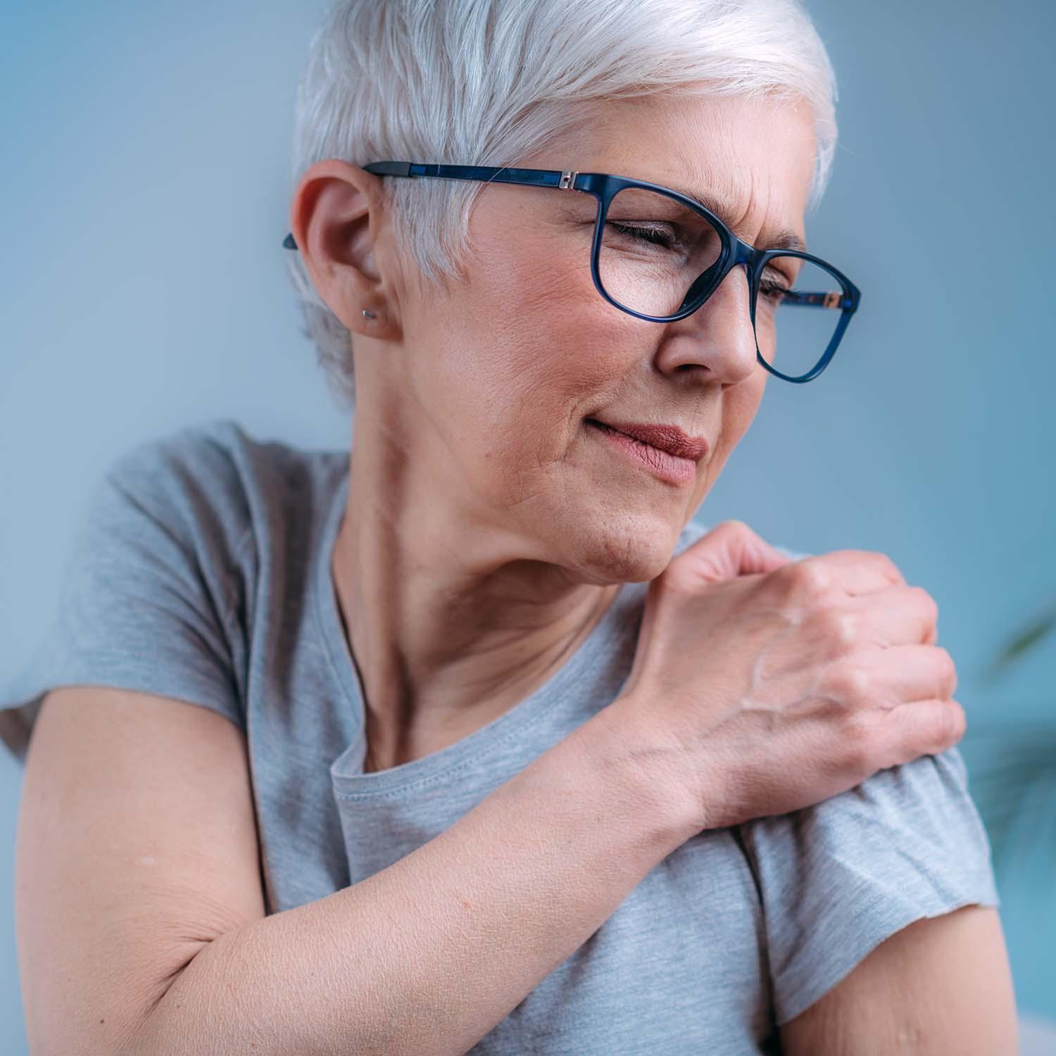 An older woman with grey hair and glasses holds her shoulder in pain.