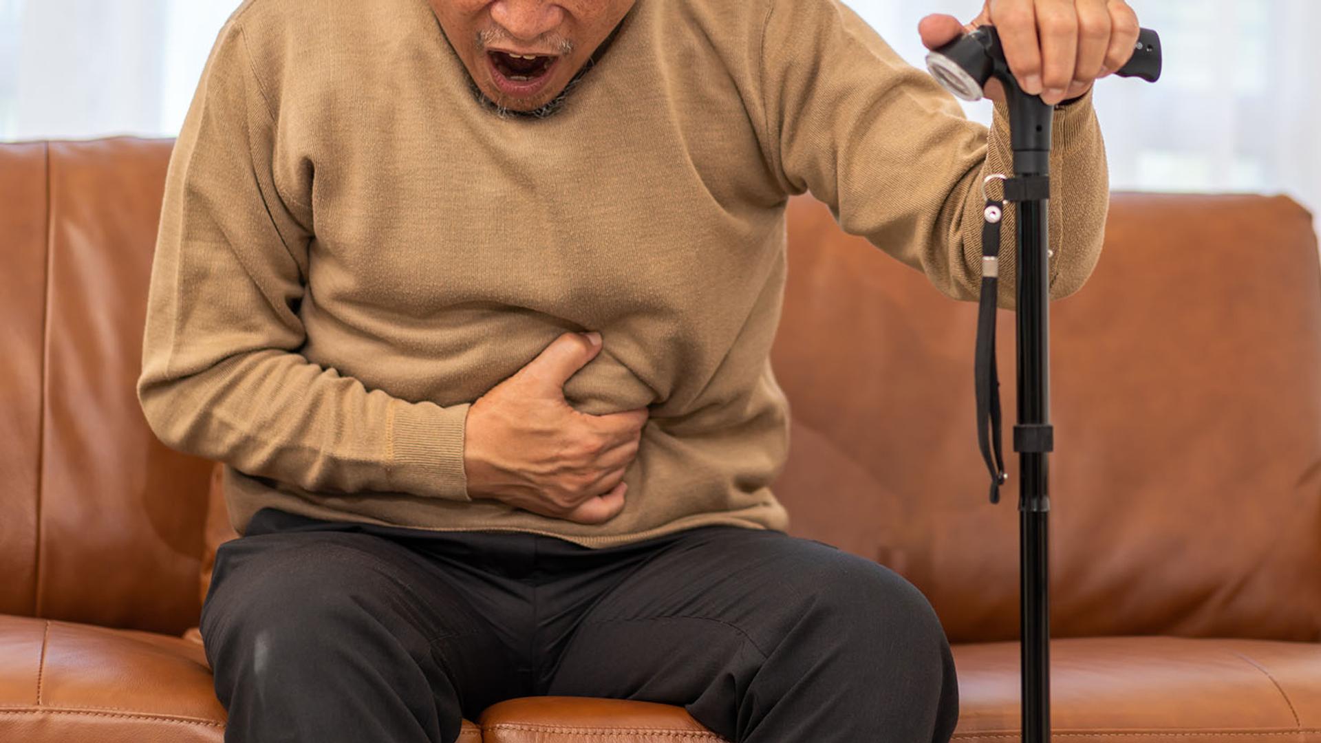 An older man sitting on a couch, clutching his stomach in pain and holding a cane.