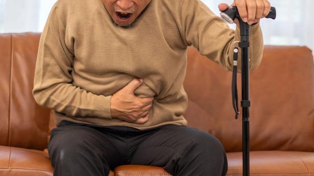 An older man sitting on a couch, clutching his stomach in pain and holding a cane.