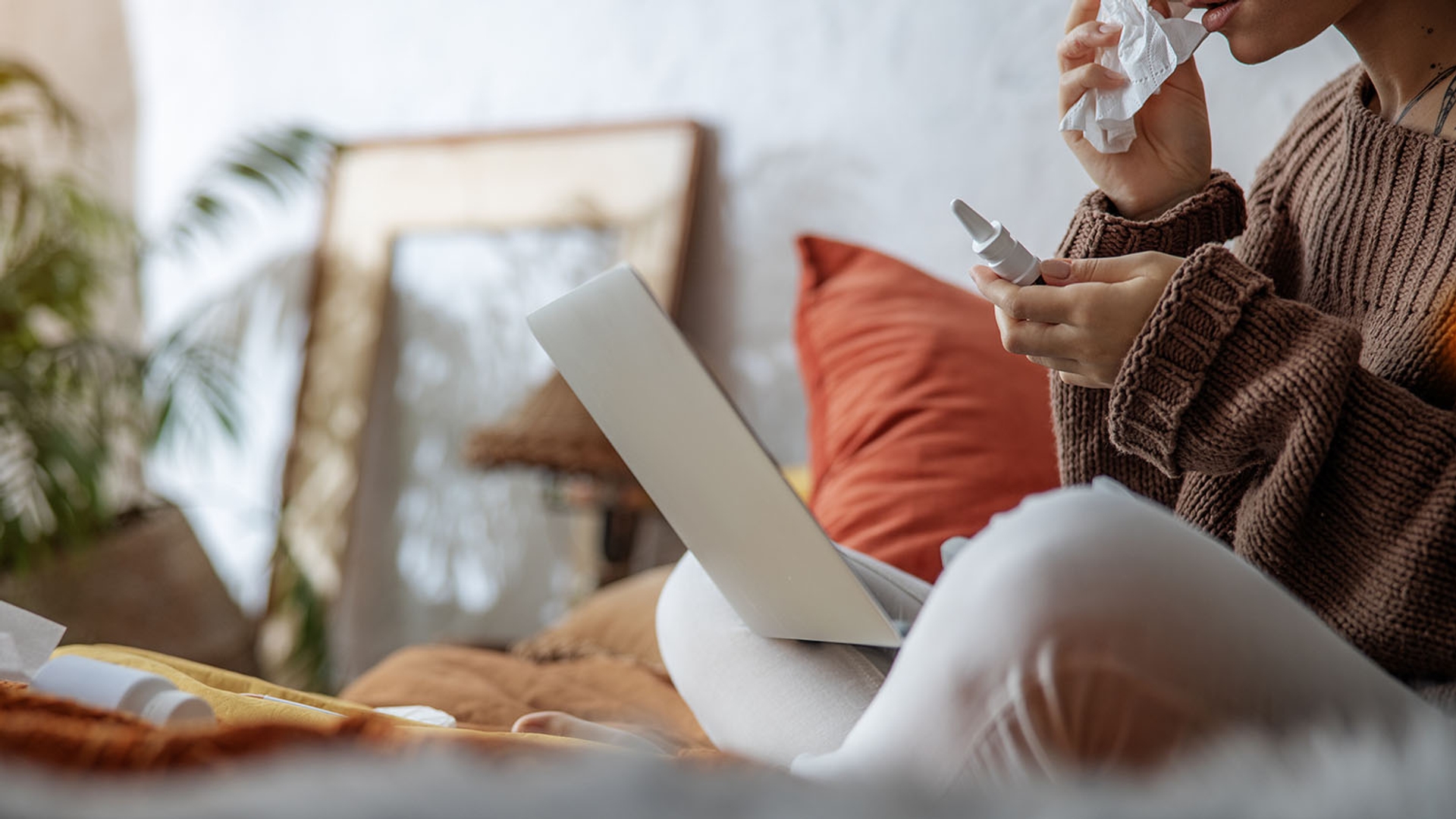 Person blowing nose with a tissue, holding nasal spray, and using a laptop.