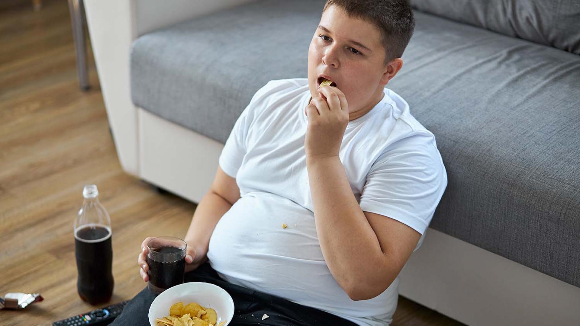 An overweight boy sits on the floor eating chips and drinking soda.