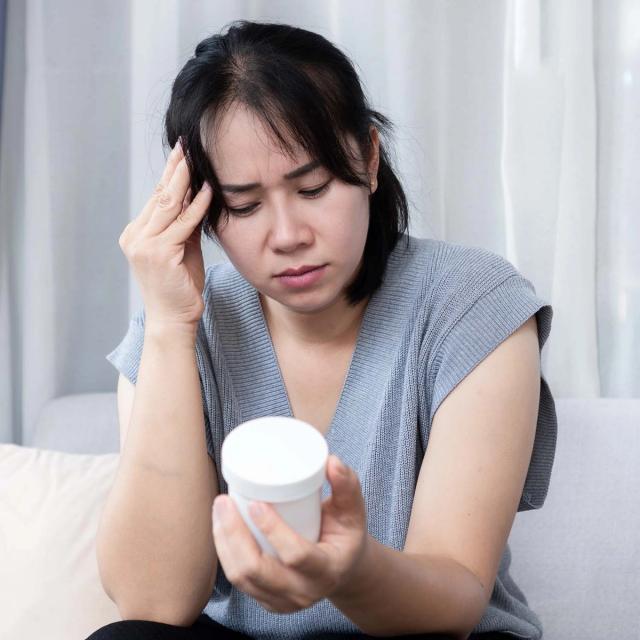 A distressed woman with a hand to her temple looks at a medicine bottle.