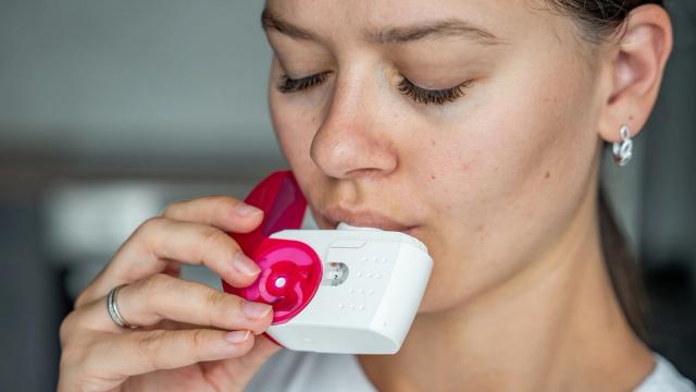 A woman with closed eyes uses a white and pink inhaler.
