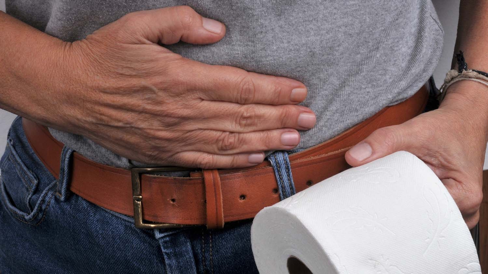 A person clutches their stomach while holding a roll of toilet paper.