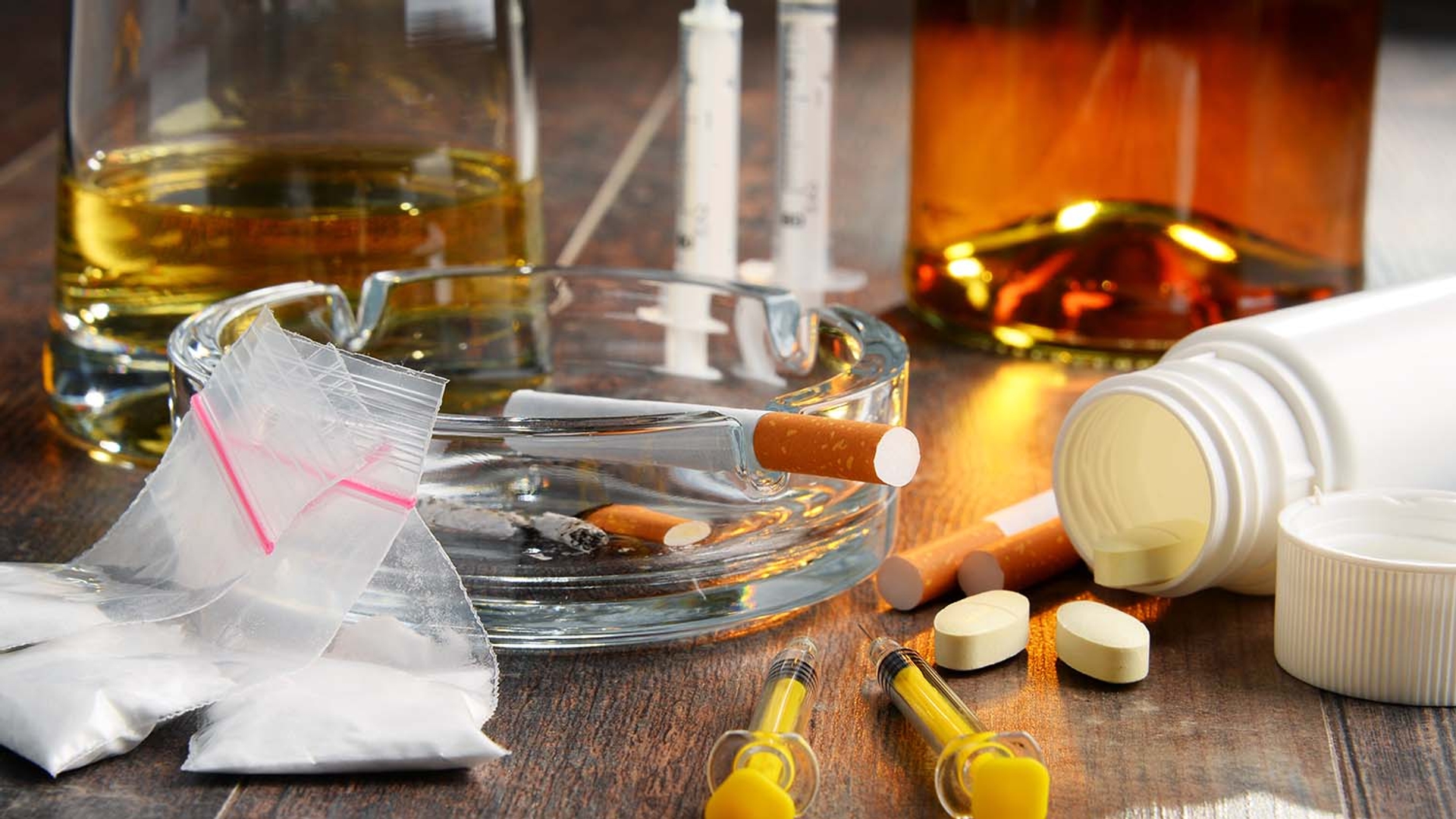 Illegal drugs, alcohol, tobacco, syringes, and prescription pills on a table.