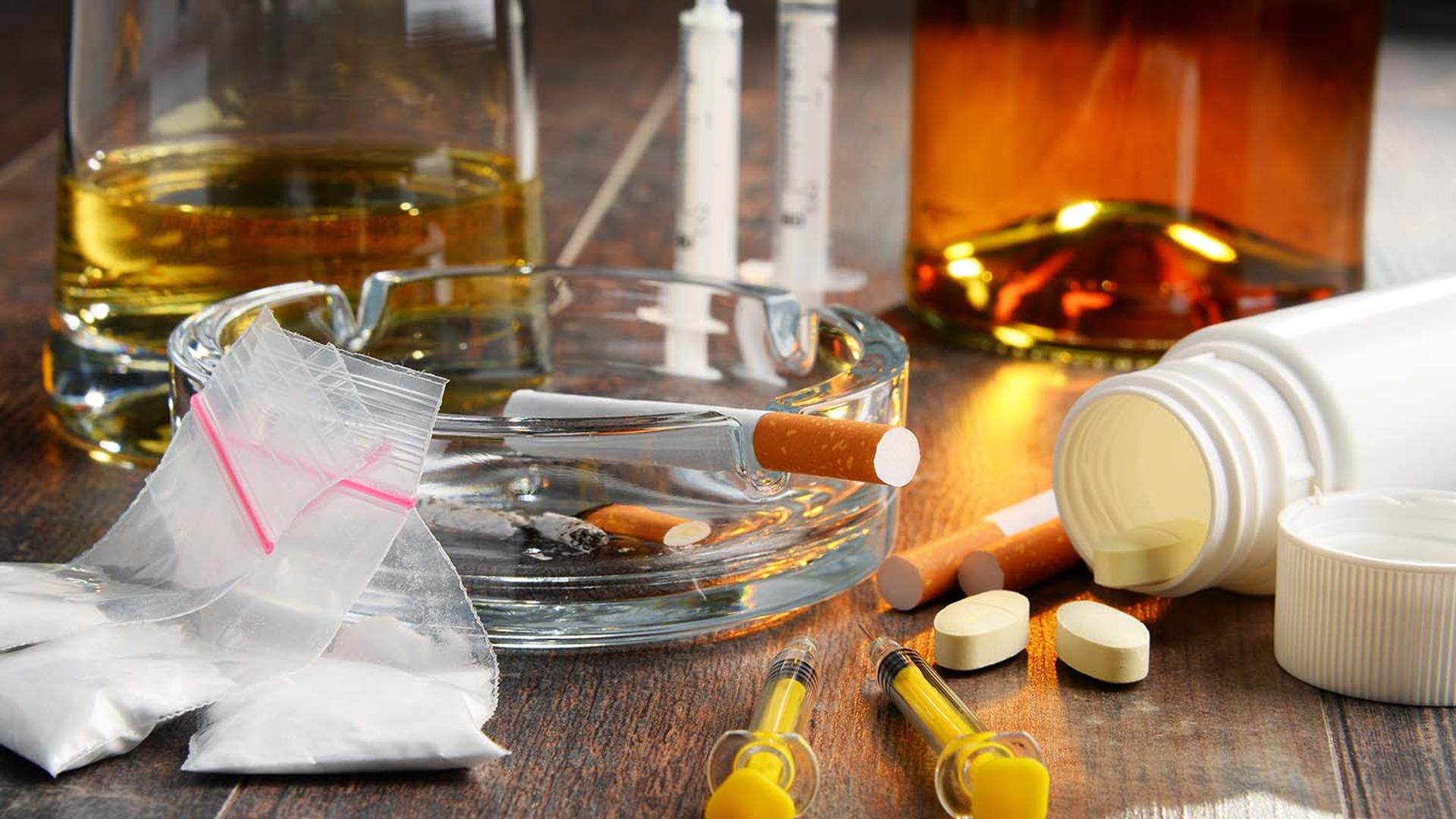 Illegal drugs, alcohol, tobacco, syringes, and prescription pills on a table.