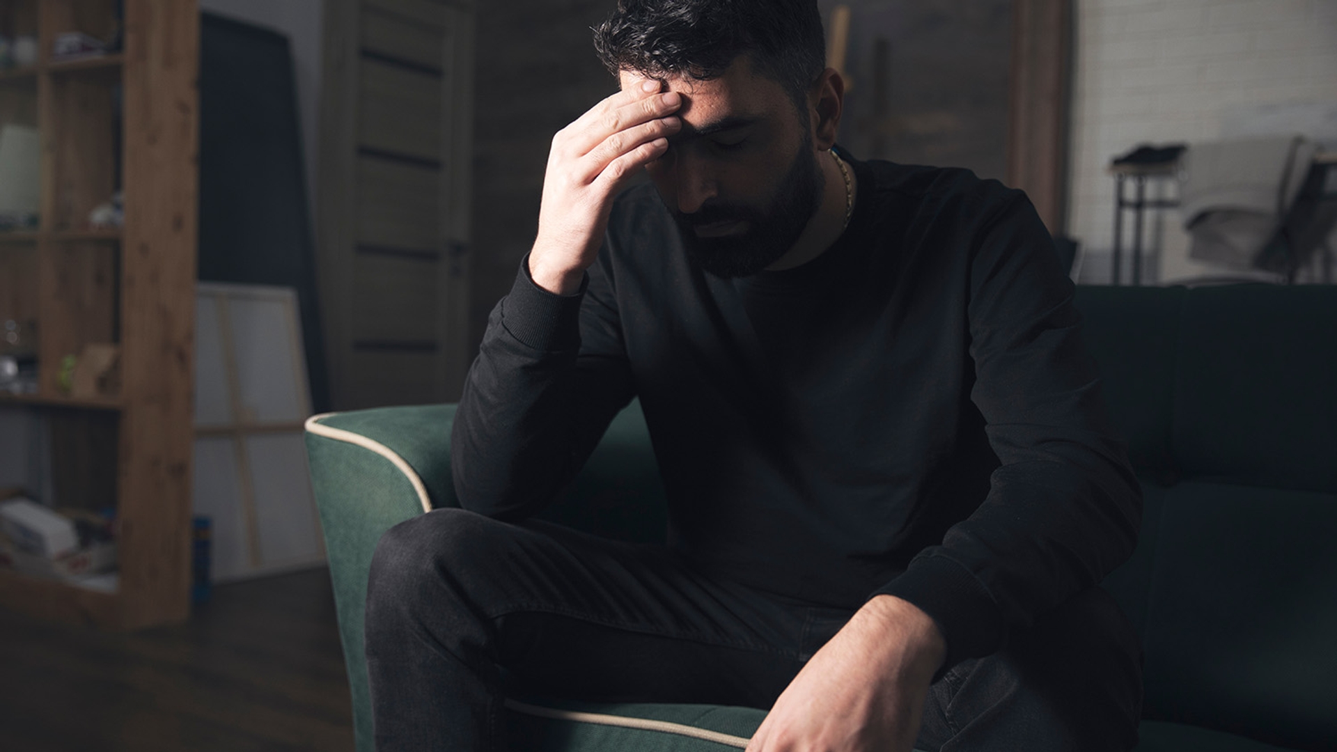 A man sits on a couch with his hand on his forehead, looking distressed.