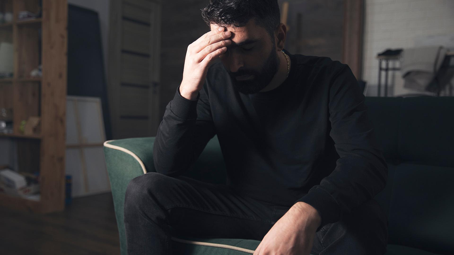 A man sits on a couch with his hand on his forehead, looking distressed.