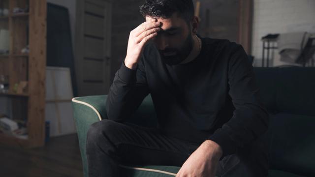 A man sits on a couch with his hand on his forehead, looking distressed.