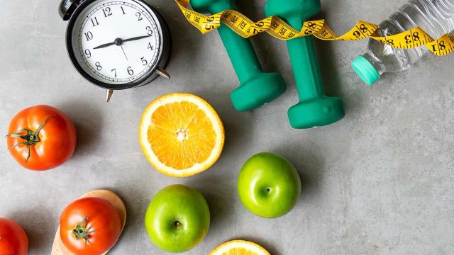 Health and fitness items including an alarm clock, dumbbells, water bottle, measuring tape, fruits, and vegetables on a grey surface.