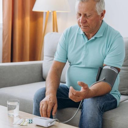 Elderly man sitting on a couch, checking his blood pressure with a digital monitor while looking at pills on the table next to a glass of water.