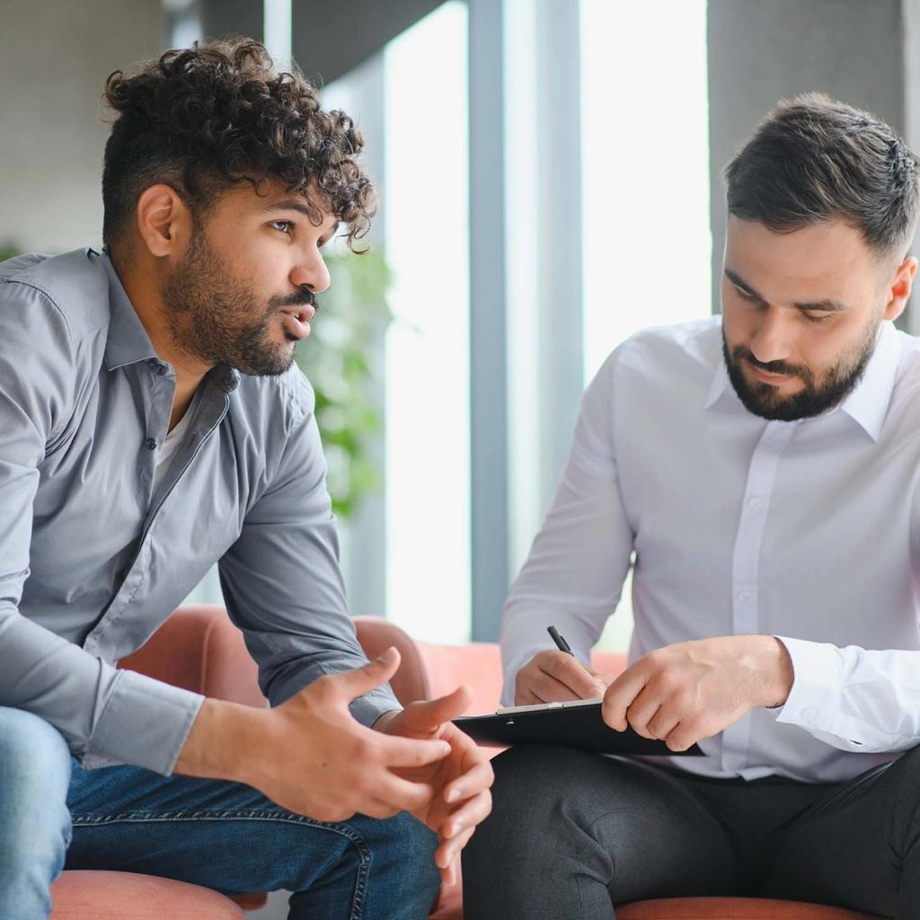 Two men in consultation; one speaks while the other takes notes on a clipboard.