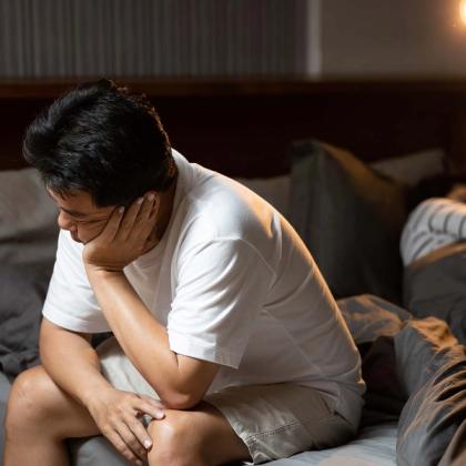 Man sitting on a bed with his head in his hand, appearing distressed or thoughtful, in a softly lit room.