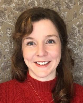 Woman with a smiling expression wearing a red sweater, with medium-length brown hair, in front of a grey ornate wallpaper background.