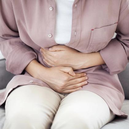 Close-up of a woman sitting with hands on stomach possibly experiencing abdominal pain or discomfort, wearing a pale pink shirt and white inner shirt.