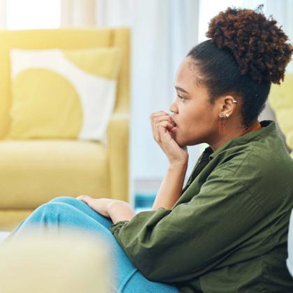 Young woman sitting thoughtfully on a couch in a brightly lit room, looking pensive