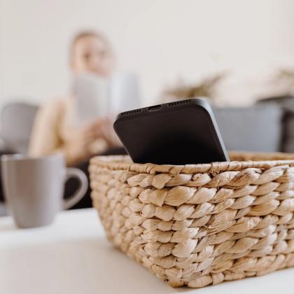 Smartphone in a wicker basket on a table with a blurred woman reading a book in the background