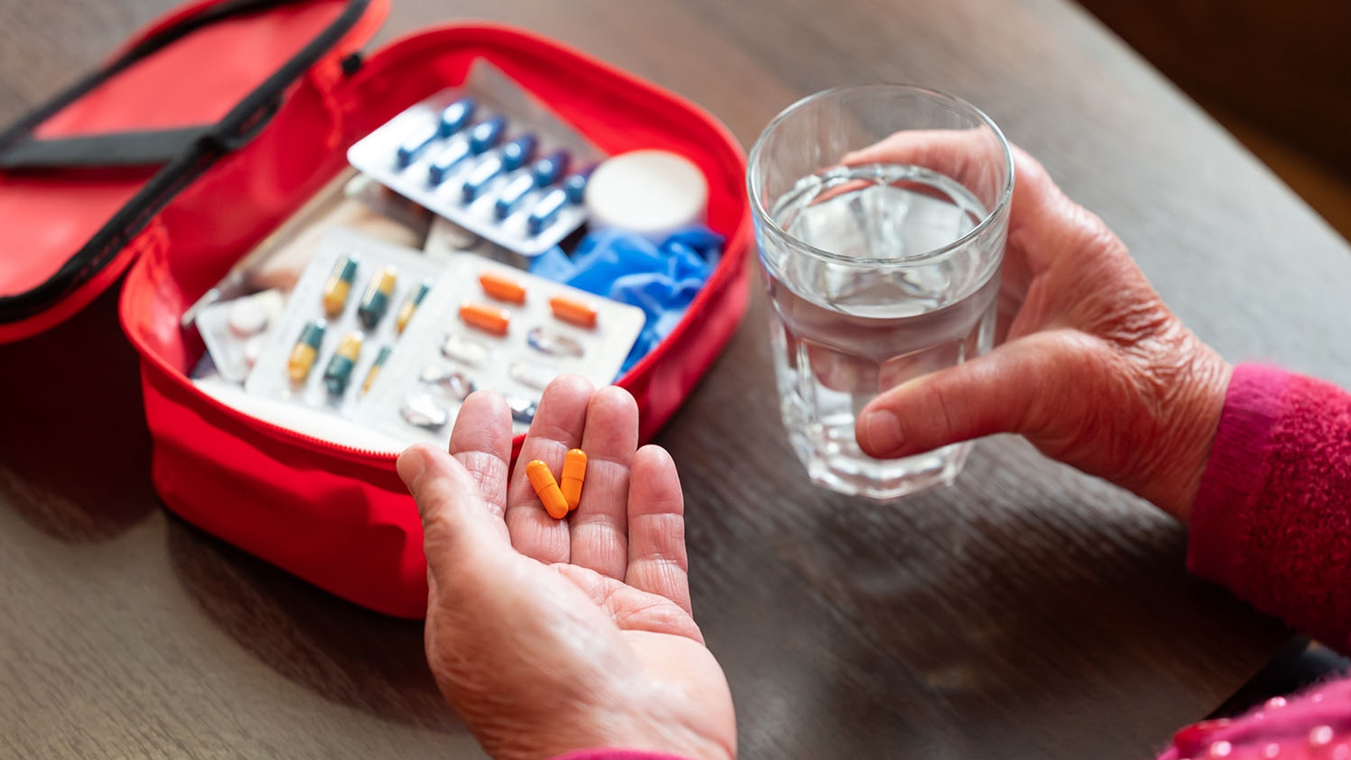 Elderly hands holding two orange pills and a glass of water, with a red first aid kit filled with medication in the background.