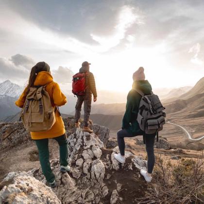 Three hikers with backpacks on a mountain summit at sunset, overlooking a scenic valley and winding road.