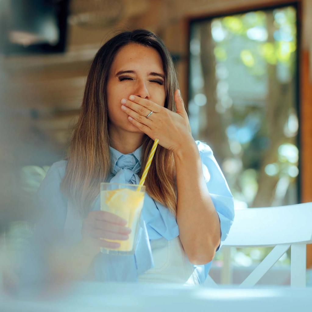 A woman grimacing with her hand over her mouth while holding a yellow drink.