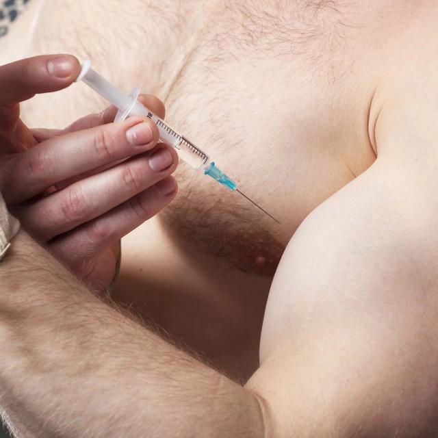 A hand holding a syringe with the needle poised to inject into a muscular upper arm.