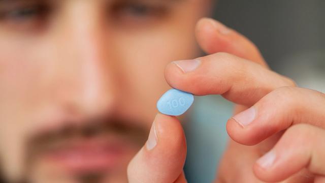 A man holds a blue, oval pill marked "100" between his fingers.